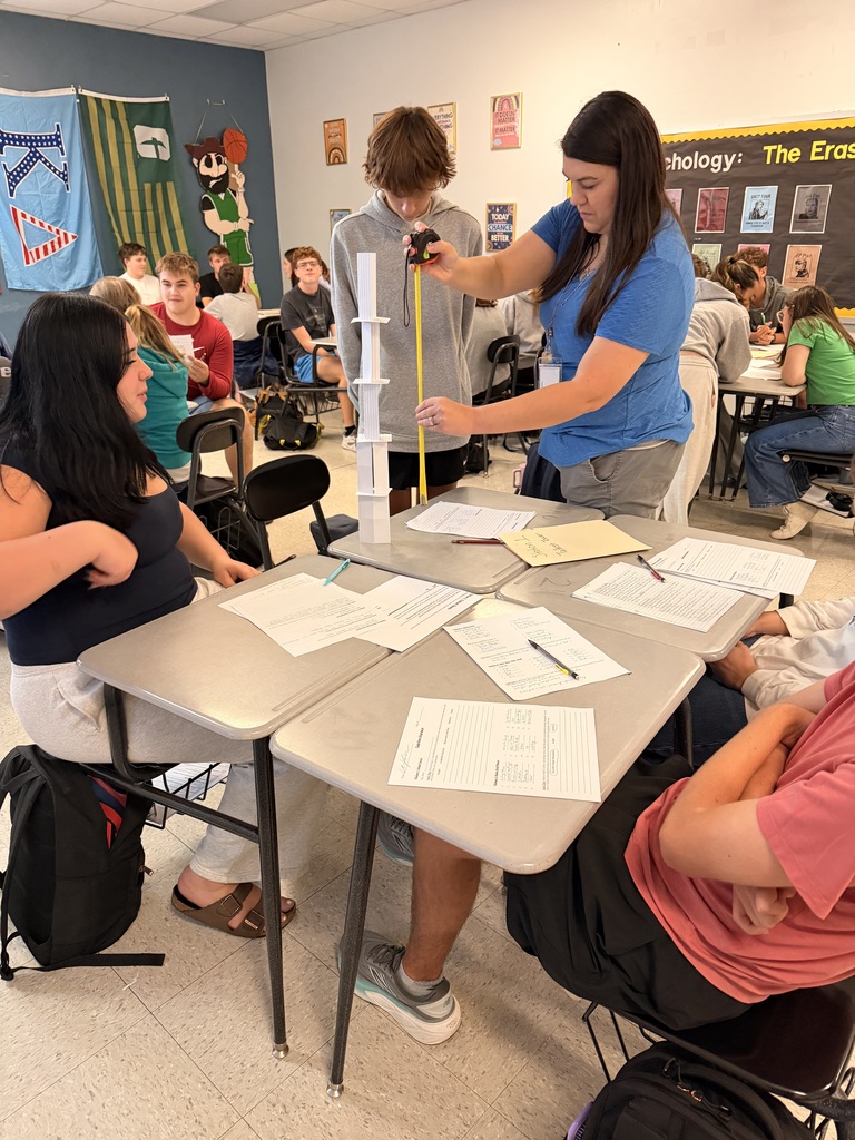 A classroom with desks. A teacher measures a tower of cups. A woman sits at a desk. Students sit at tables.