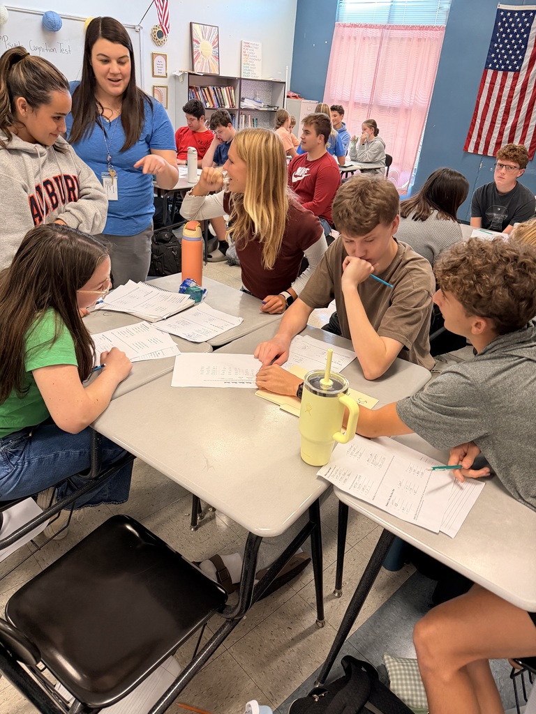 A classroom with students seated at tables, discussing papers. A teacher stands next to one of the tables.