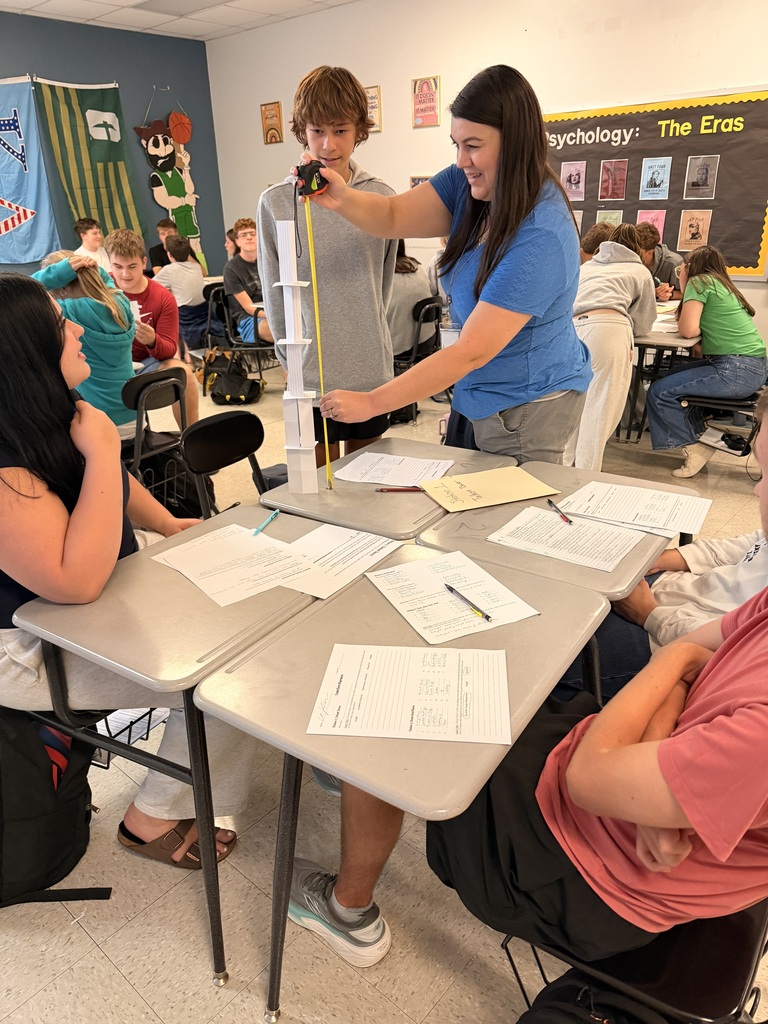Several students in a classroom, one measuring a tower of cups on a table with papers.