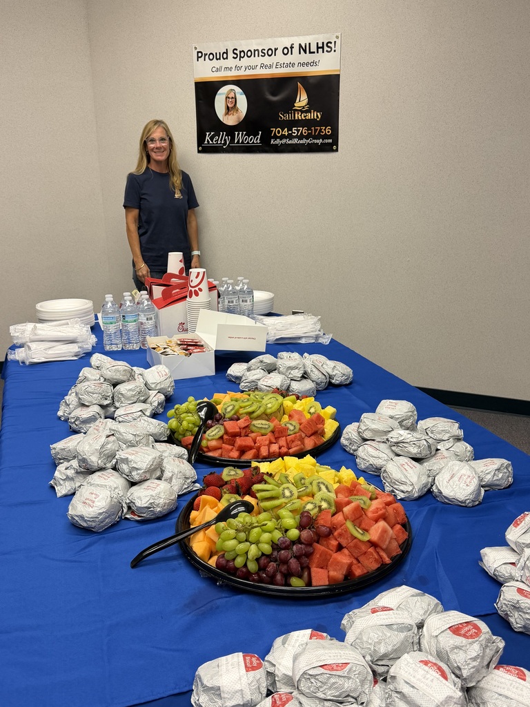 A table with blue cloth has two trays of fruits and multiple plastic bags. A woman stands near the table. A sign on the wall reads "Proud Sponsor of NLMK."