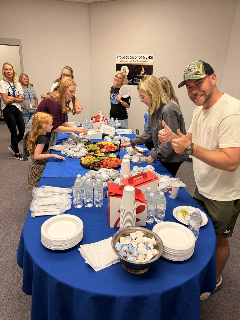 A group of people standing around a table with food, plates, cups, and a bowl of candy.