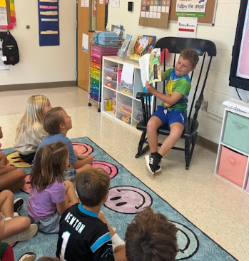 A group of children sit on a carpet, one reading a book while others listen. Shelves with books and classroom items are in the background.