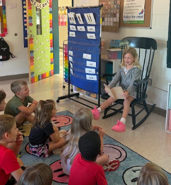 Several children sit on a rug with smiley faces while a girl reads in a rocking chair.