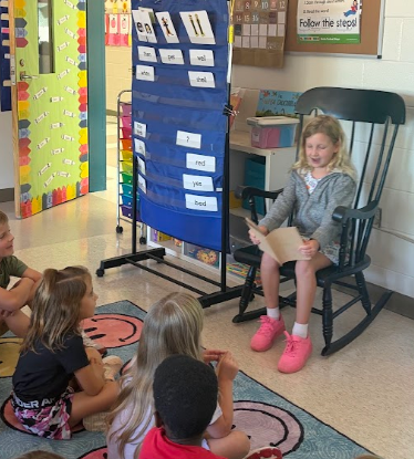 Children seated around a teacher on a rocking chair in a classroom with a word wall.