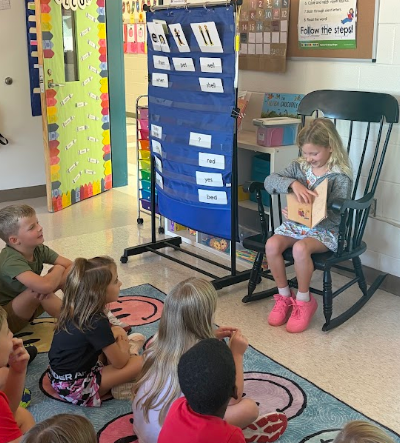 A classroom with children seated around, one girl reads a book in a chair. A blue educational board stands nearby.