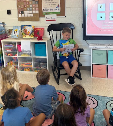 A group of young children sit on a rug while a boy reads aloud from a book.