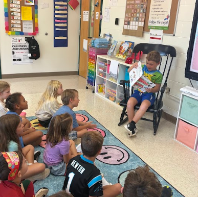 A classroom with students seated on a rug. One boy reads aloud from a book.