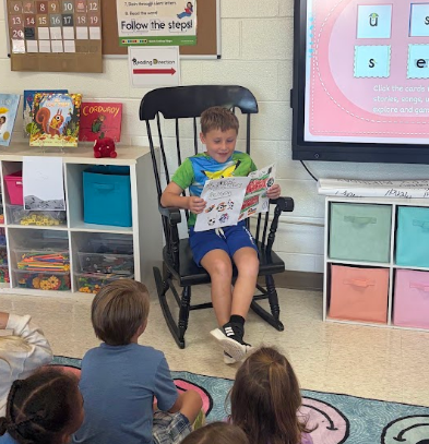 Children seated in chairs with one boy reading from a book. A monitor behind displays a pink screen.