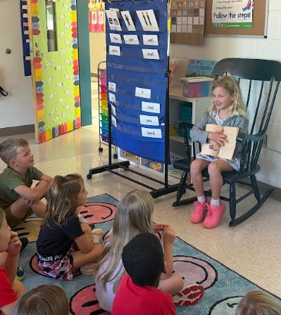 Children sitting on the floor and a girl in a rocking chair. A blue board is mounted on a stand.