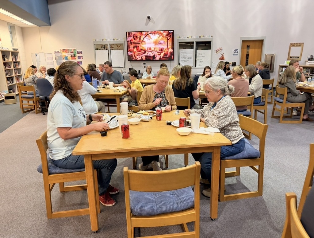 People seated around tables in a dining area. A woman in white stands at a table with food.