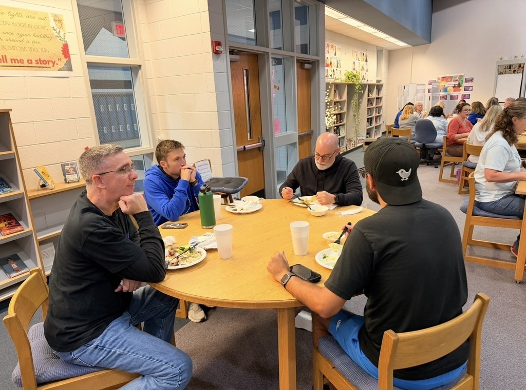 Five men seated around a table in a library. Each has plates of food and beverages. A bookshelf is in the background.