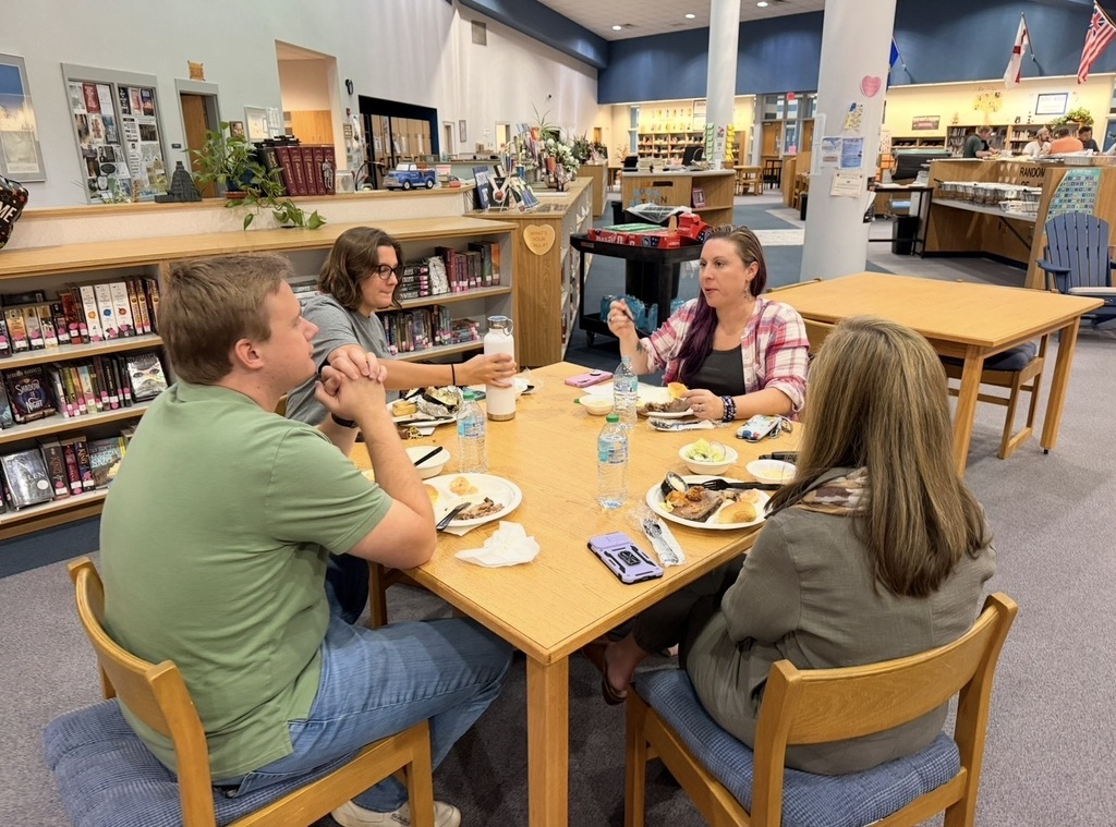 Four individuals sit at a table in a library with food, books, and a pillar behind them.