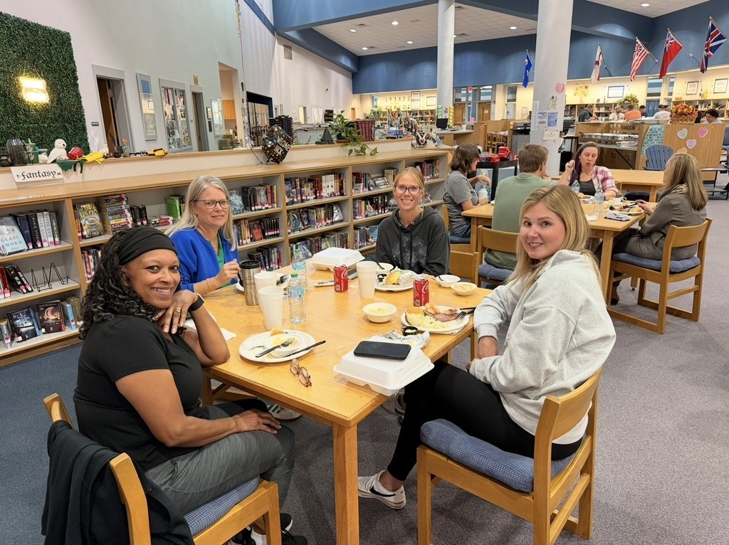 Six people are sitting at a table in a library, smiling and talking. One of them is holding a cell phone.
