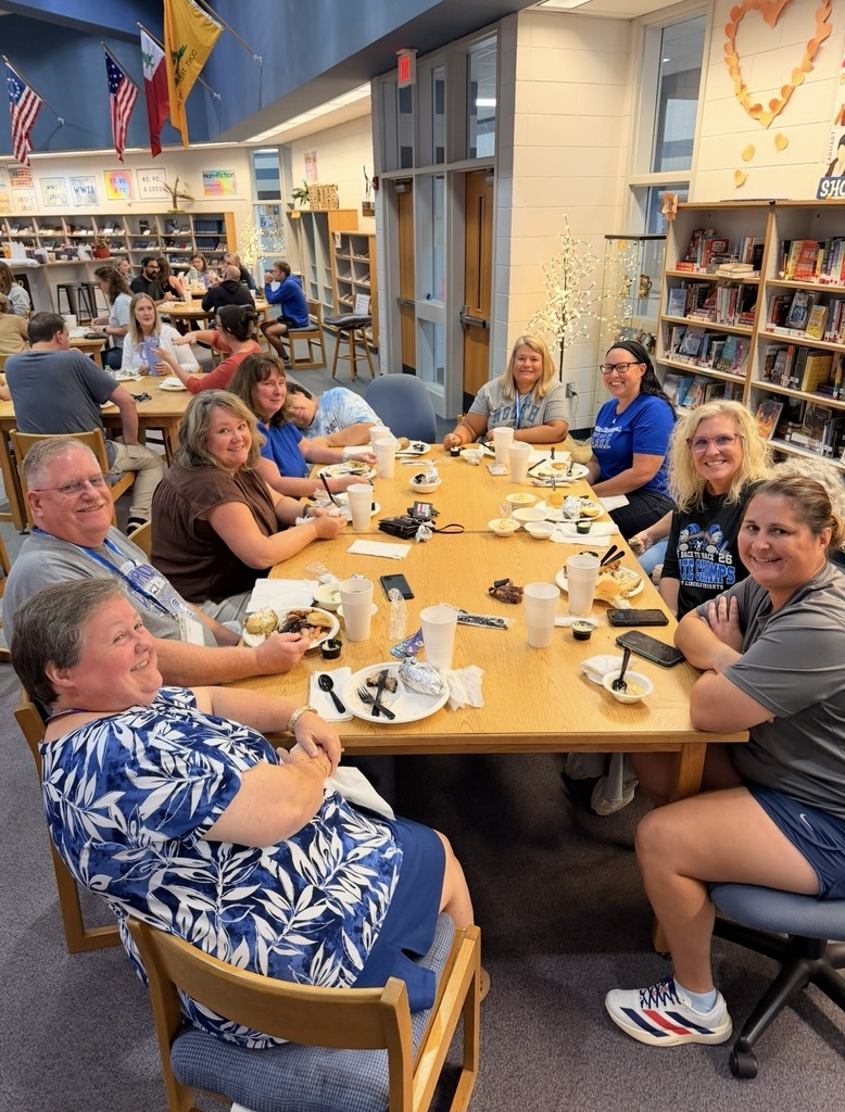 A group of people at a long table in a library. Some are seated, others standing, all holding food and drinks.