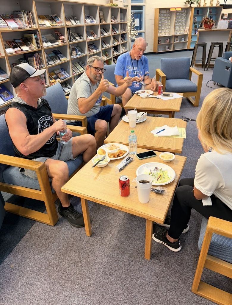 Four people in a library meeting room, seated around tables with food and drinks. A bookshelf is behind them.