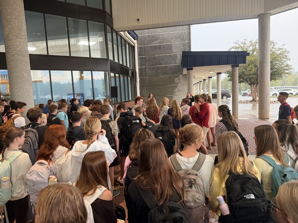A crowd of people, carrying backpacks, gathers in a lobby. Glass windows, a tree, and distant vehicles are visible.
