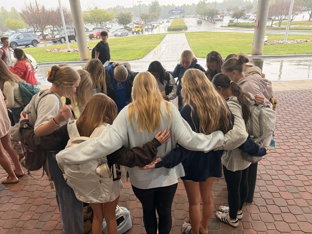 A group of people stand in a circle on a brick floor, some wearing backpacks. They face outward, creating a circle, in a wet area.