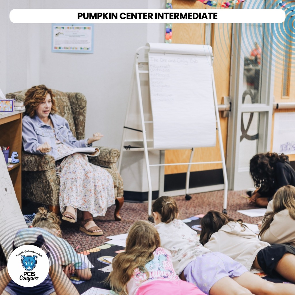A woman sits in a chair reading to students on a rug. A whiteboard stands behind her.
