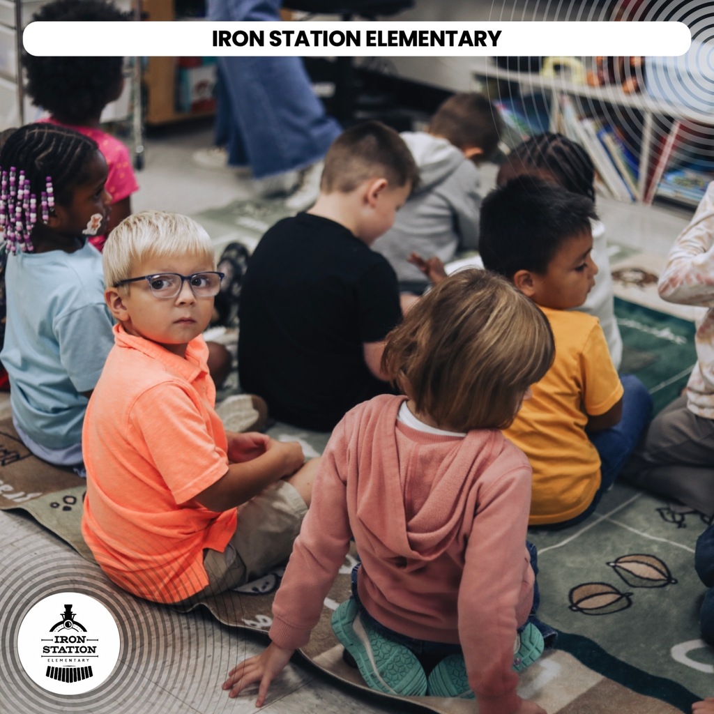 A group of children sitting on the floor, engaged in classroom activity, with a rug and shelves in the background.