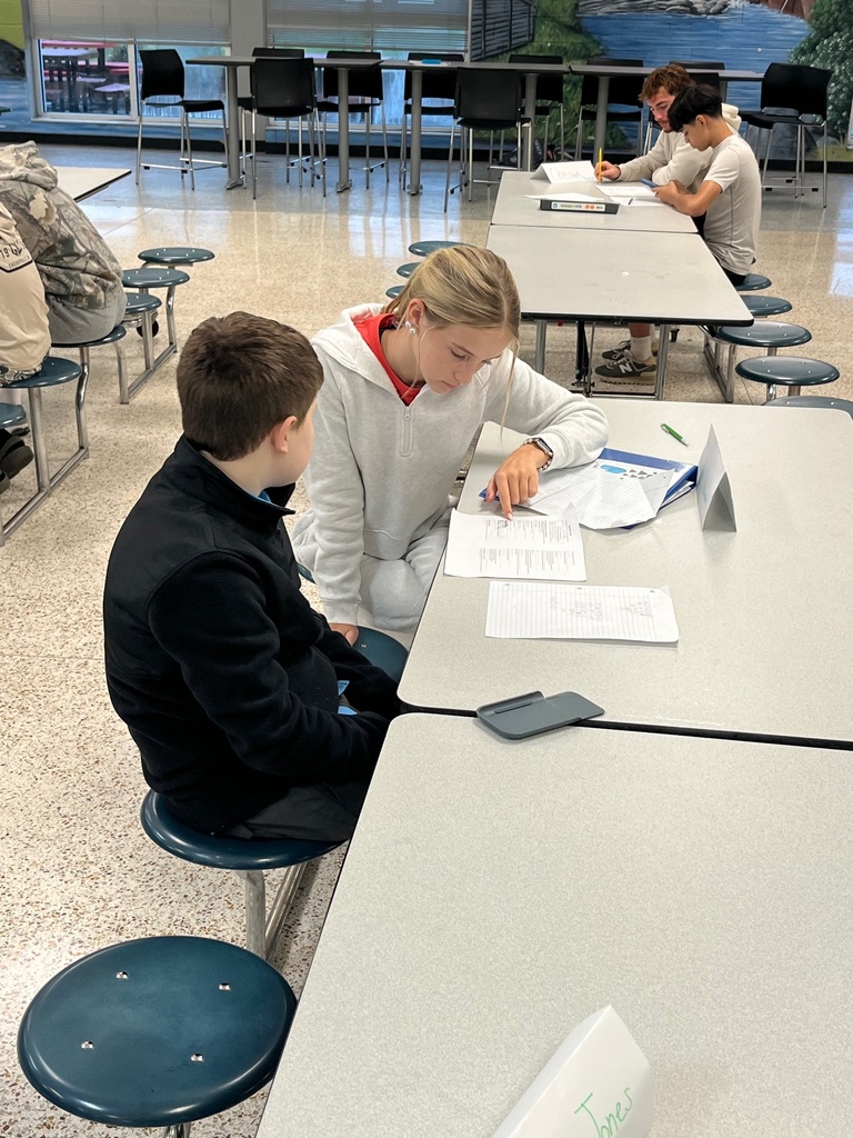 Three people sit at tables in a room with a table and chairs in the background. Two individuals converse, while the third writes.
