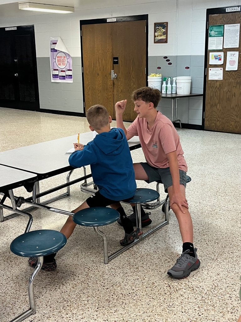 Two boys sitting at a cafeteria table. One has a pencil and is seated while the other stands with hands on knees.