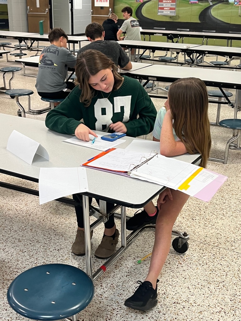 Two students sit at a desk in a cafeteria, one writing on paper while the other looks on.