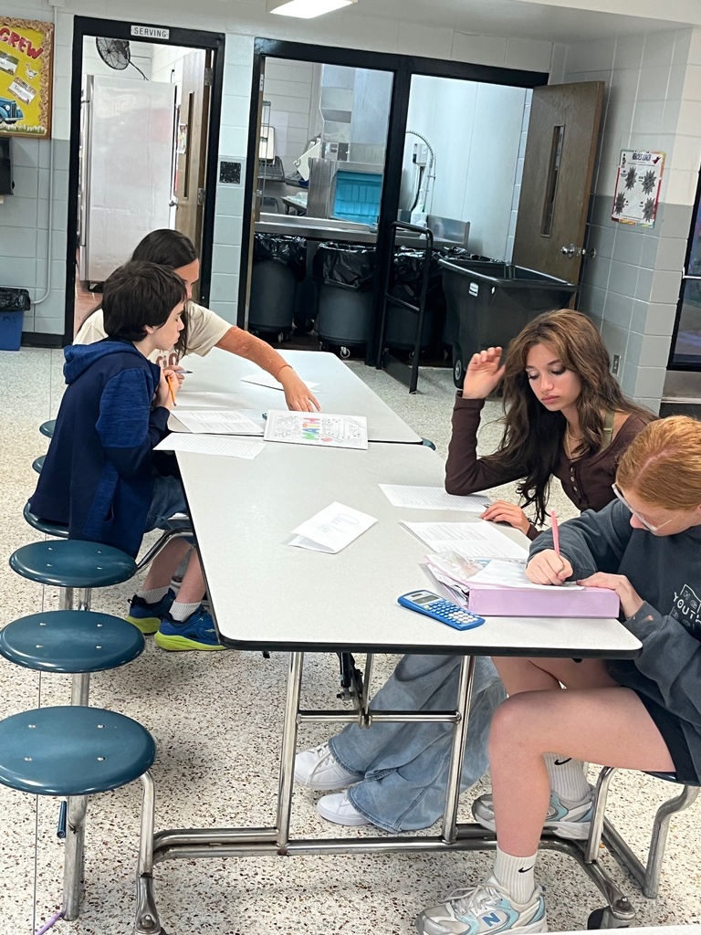Four people seated around a table in a classroom, writing in notebooks and using pens.
