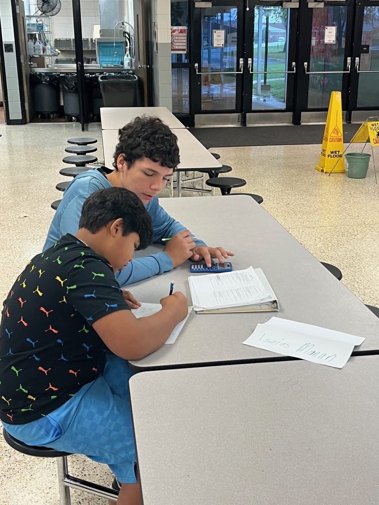 Two young males seated at a table, one holding a pen and paper, the other holding a tablet.