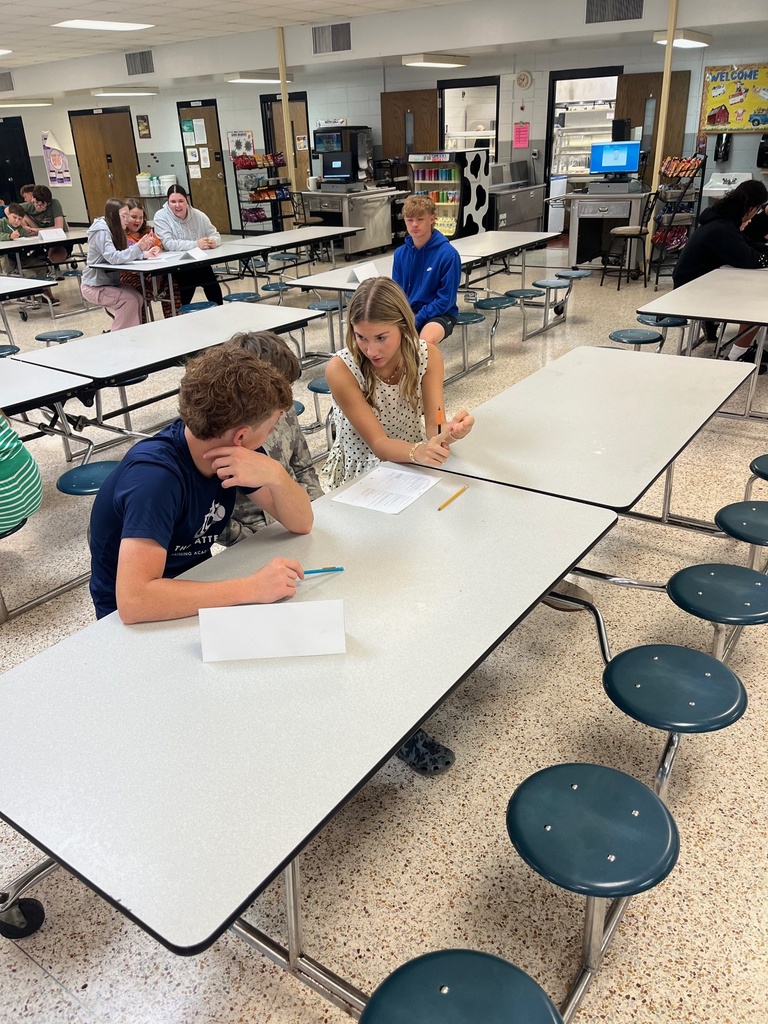 Group of students in a cafeteria. Two students sit at a table, one with head down, other with pen.