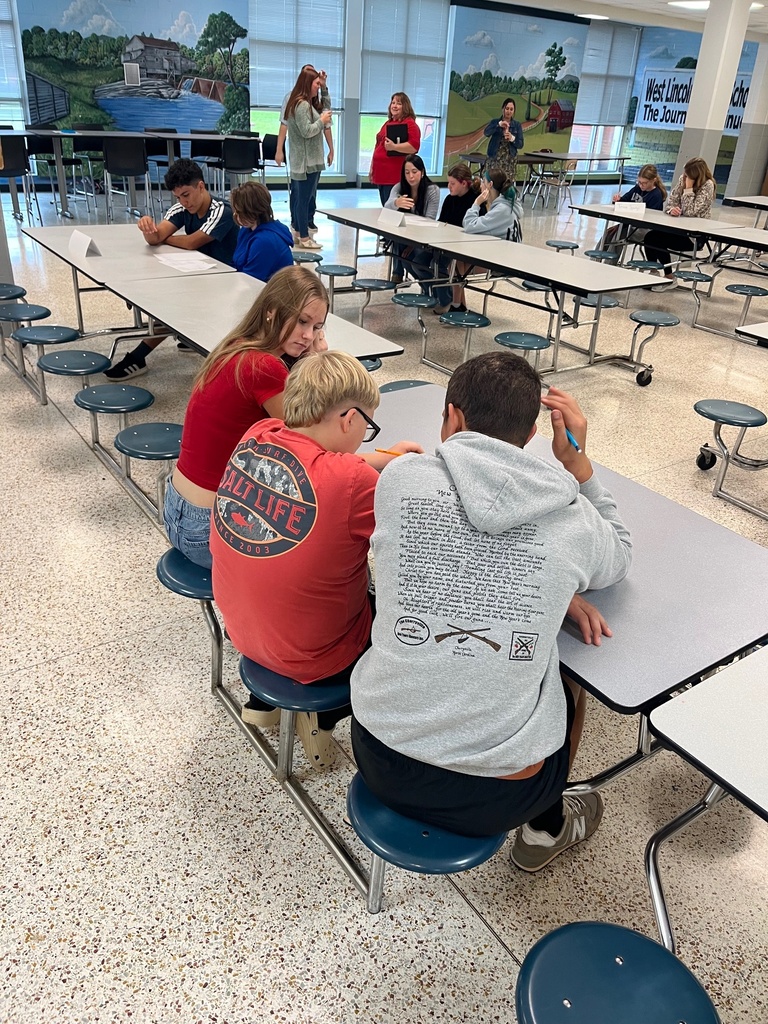 Students sit around tables in a cafeteria. They are seated at long tables with chairs, some of which have blue seats.
