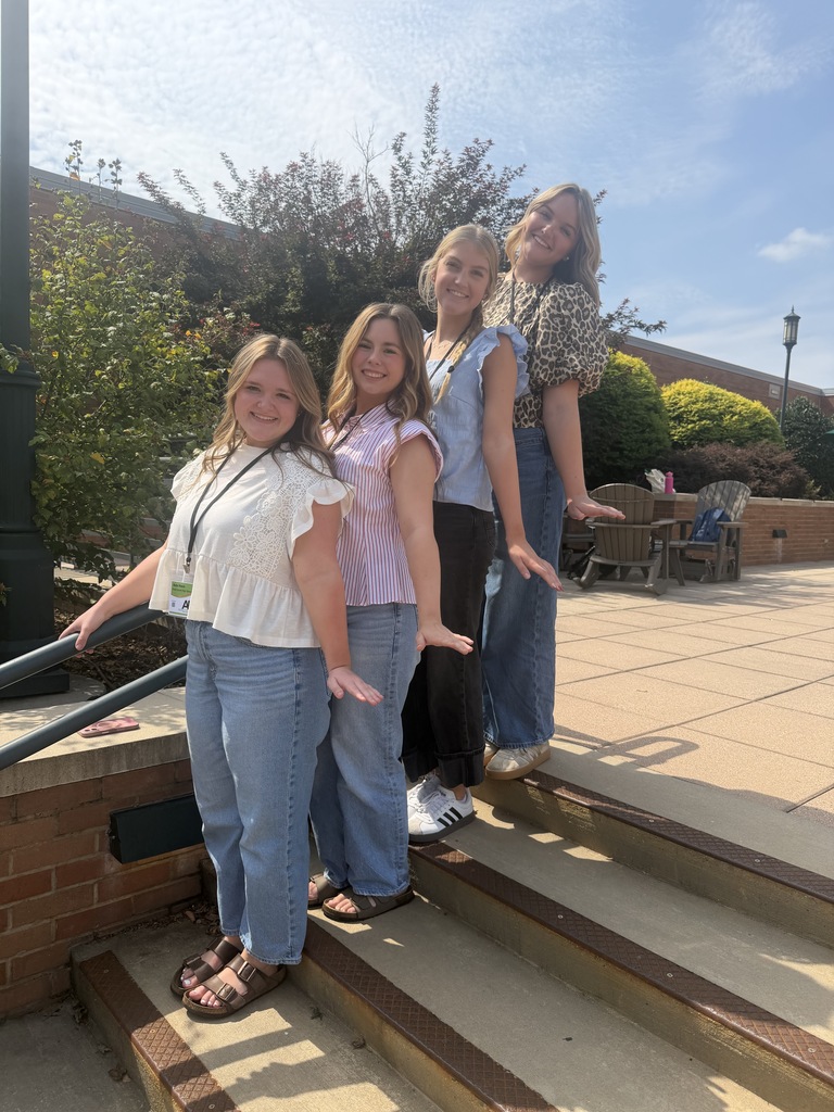 Four young women pose on outdoor stairs, wearing casual clothes. Behind them are plants and a brick wall.