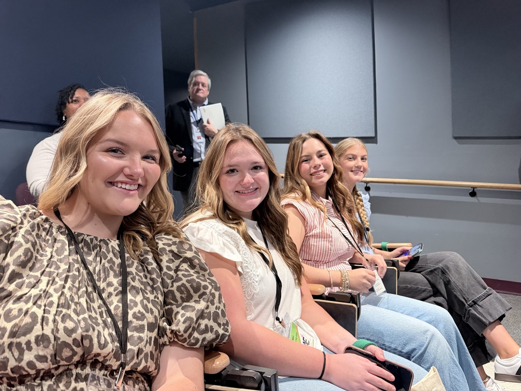 A group of four young women sitting in chairs, smiling. One wears a leopard-print blouse.