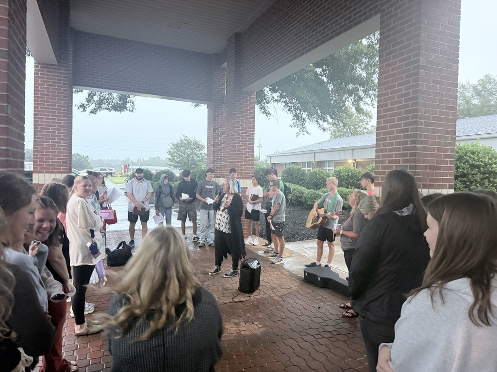 People gather outside a brick building, with a few holding instruments and others standing. Trees and distant buildings are visible.