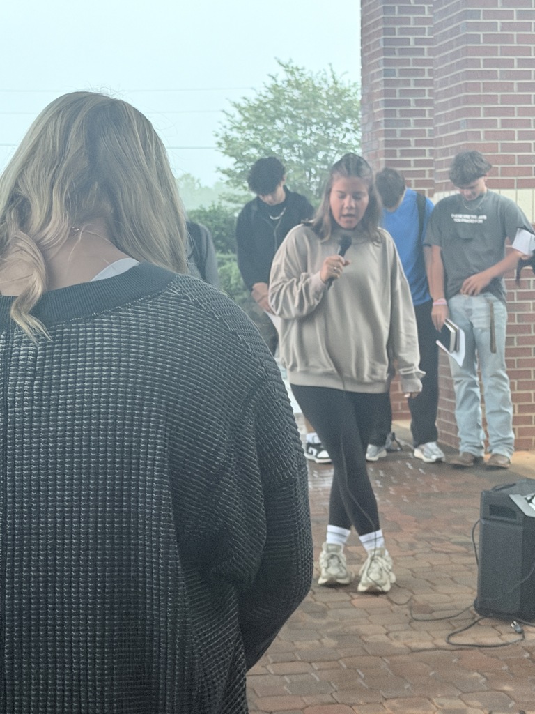 A young person holds a microphone and speaks at an outdoor gathering. Several others stand behind her.