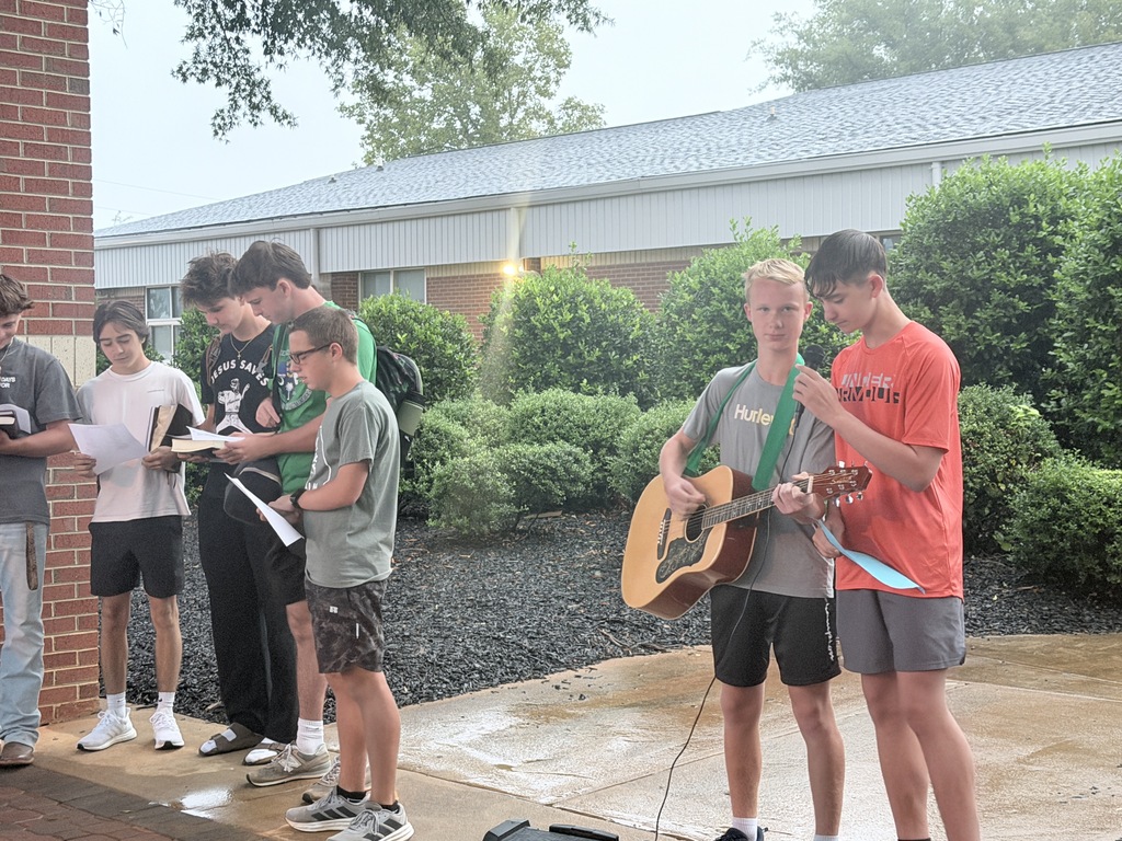 Seven teens are standing outside a brick building, with one holding a guitar and singing while others stand around him.