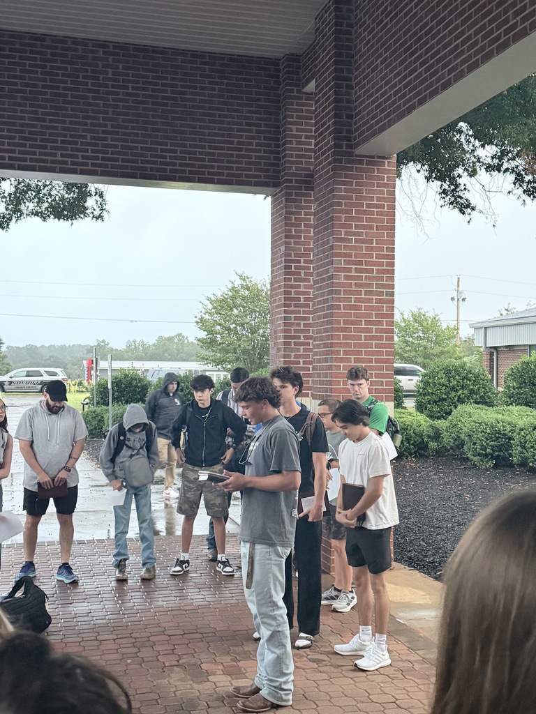 Several people gather under a brick structure, looking at a man standing. Trees, shrubs, and a car are in the background.