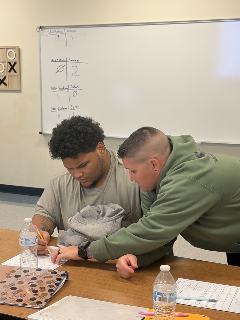 Two individuals, one in green and the other in gray, are bent over a table, reviewing papers.