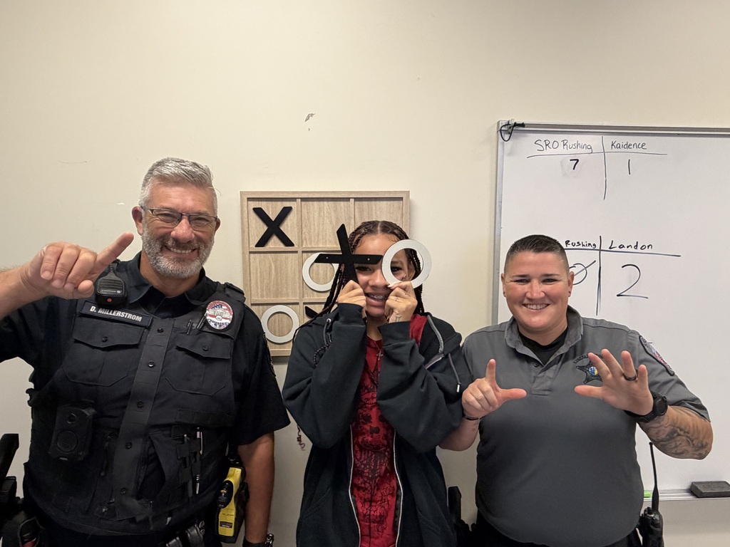 Three people posing for a photo. One holds a pair of scissors. Behind them, a whiteboard has numbers and letters on it.