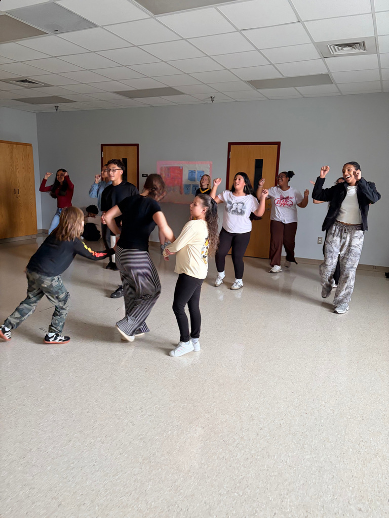 Group of people, in various attire, dancing in an indoor space with white walls and doors.