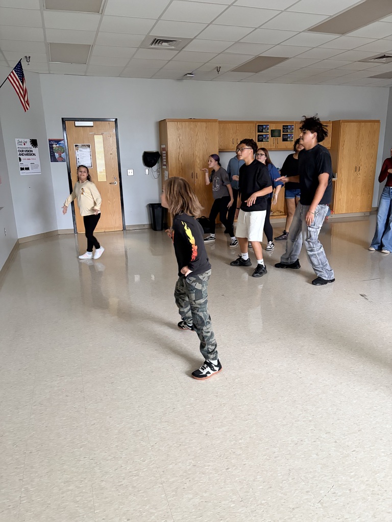 People stand in a classroom, some in sneakers, with one person in front wearing a black shirt.