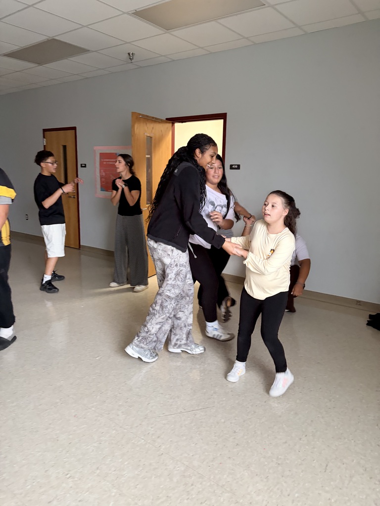 A group of people in a white room with open doors. One woman and a young girl dance.
