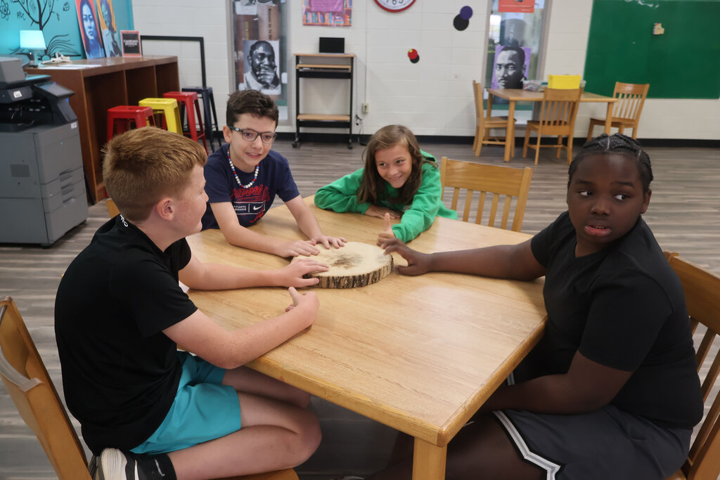 Four children seated around a table, with a boy playing a drum in the center. The room has chairs, a table, and a green board.