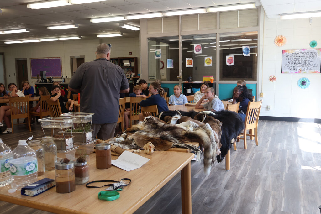 A classroom with students seated around tables. A teacher stands in front of a table with various items.