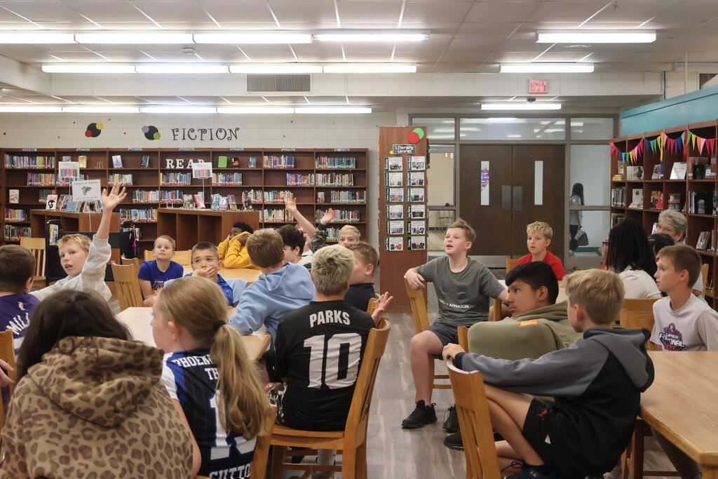A group of children sit around a table in a library, some raising hands, some sitting, and some wearing jerseys.
