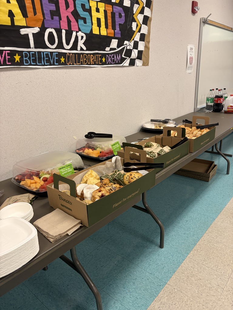 A long table holds trays with various food items. White plates are stacked at the left end. A colorful banner says "Leadership Tour."