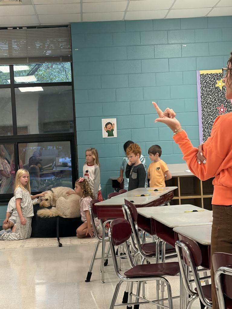 An adult in orange gestures while children gather around a large dog in a classroom.
