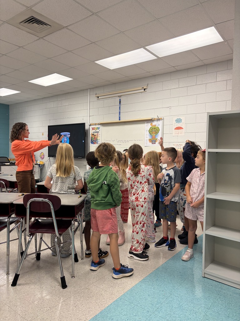 A woman stands in front of a classroom with children gathered around. A whiteboard is on the wall.