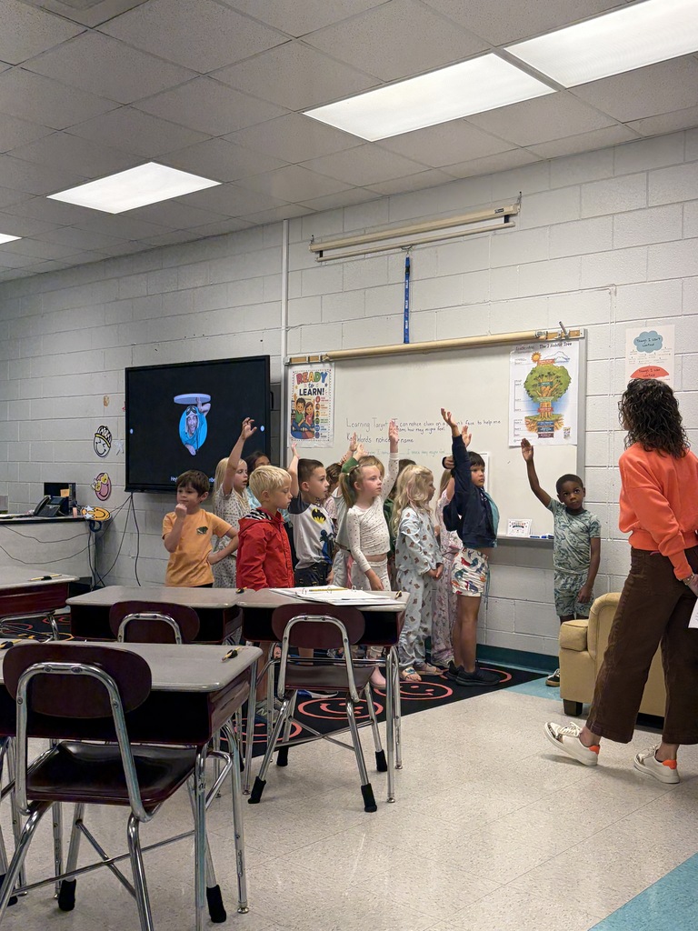 A group of children in a classroom, raising hands, facing a teacher and a whiteboard.