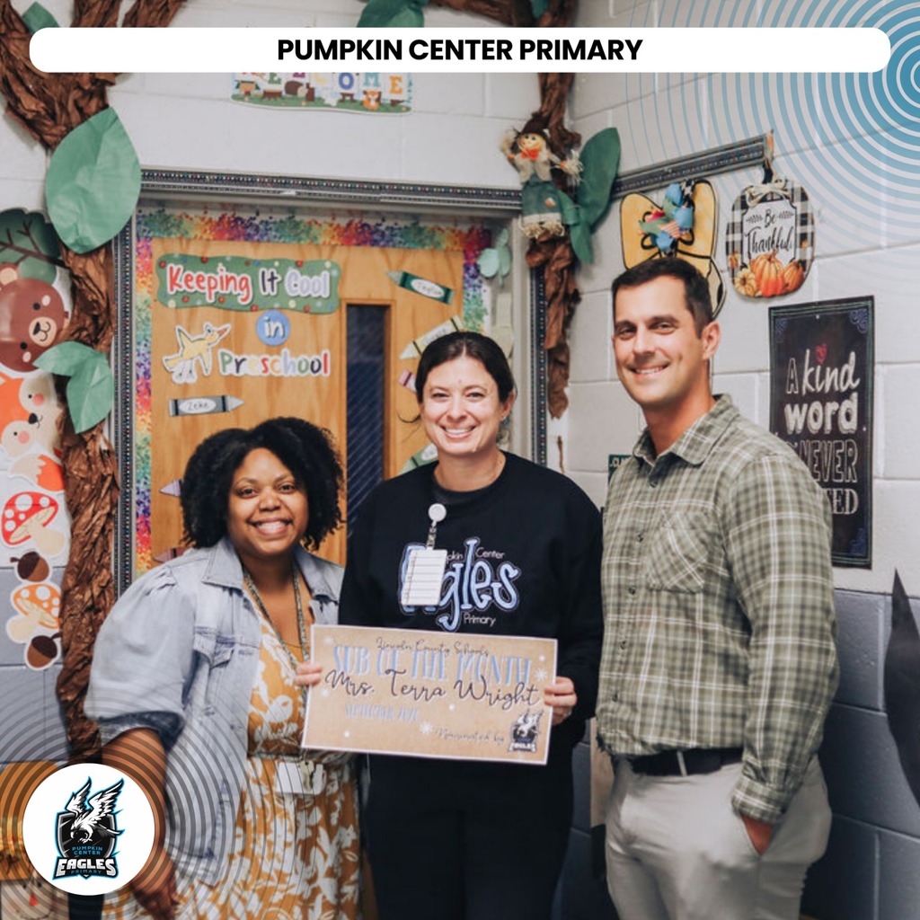 Three people stand in front of a door with "PUMPKIN CENTER PRIMARY" text, holding a sign.