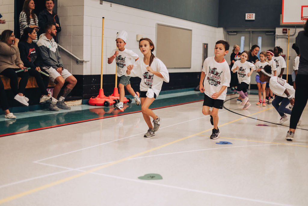 A group of children run in an indoor gym, marked by white lines on the floor. Some wear white shirts.